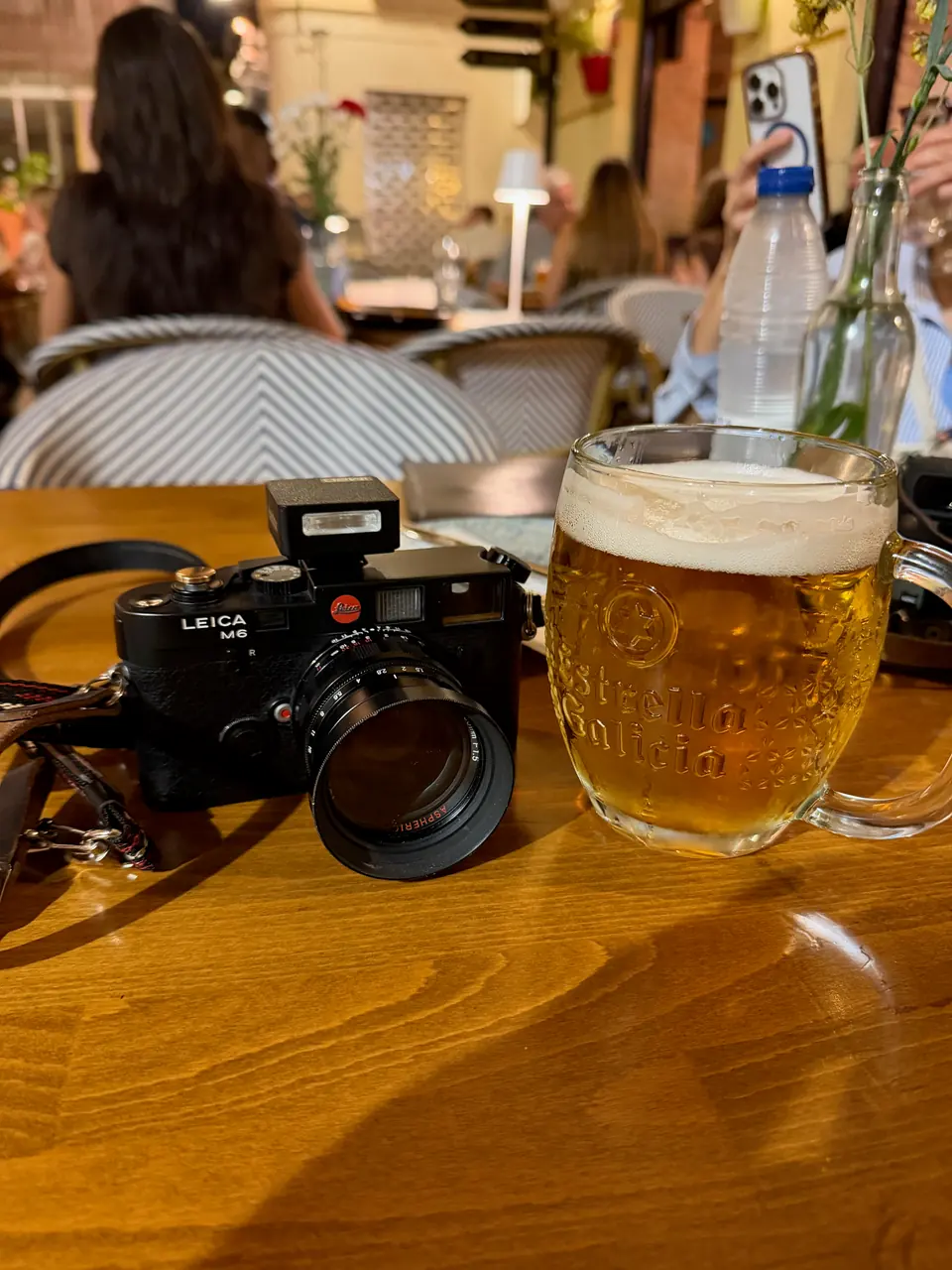 Black Leica M6 with a large black lens and a small flash in the hot shoe, its strap trailing off to the left, on a wooden café table beside a frothy mug of Estrella Galicia